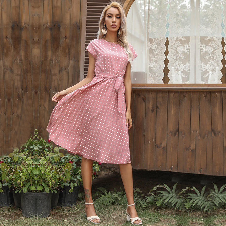 Woman in a pink polka dot dress standing outdoors with plants and a wooden wall in the background
