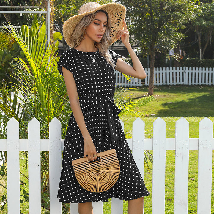 Woman in a black polka dot dress and straw hat standing in front of a white picket fence.