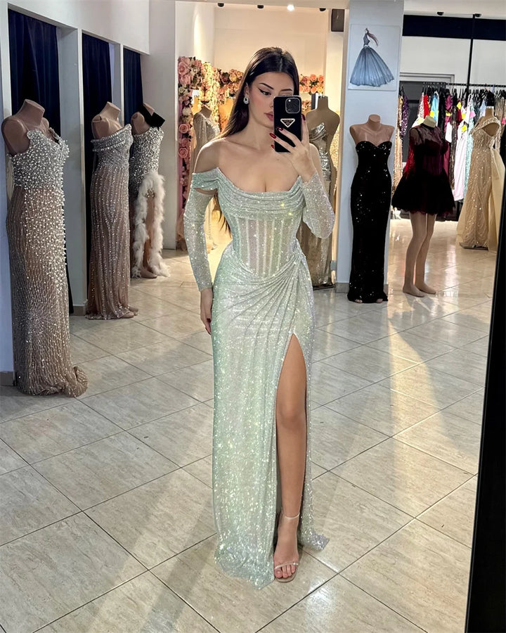 Woman in a sparkling green evening gown with long sleeves and a high slit, standing in a store with mannequins and dresses.