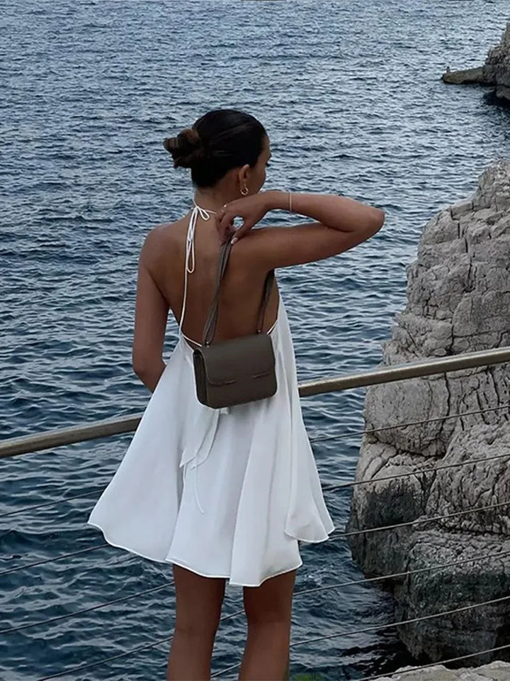 Woman in a white dress standing by the water with a gray bag.